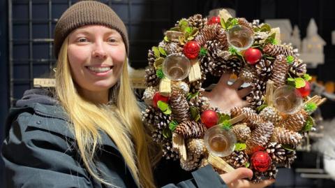 A stall holder with long blonde, wearing a brown hat and a navy jacket. She is holding a multi-coloured Christmas wreath. 