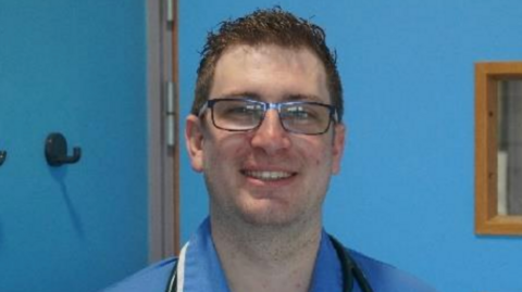 A man in a nursing uniform smiles at the camera. His uniform is blue and there is a stethescope around his neck. He has short brown hair and wears rectangular spectacles. The room his is standing in is painted bright blue.