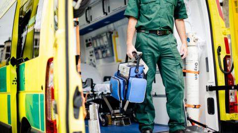 A stock image of a male paramedics in green scrubs, carrying a blue bag containing medical equipment. He is standing in the back of an ambulance with the rear doors wide open.