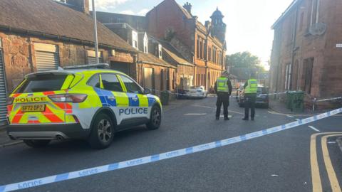 Police tape blocks off a section of a street. A police car is parked on the road and two police officers stand by the tape.