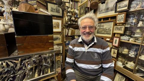 A man with grey hair is wearing a stripped brown, white and grey shirt. He is wearing a pair of glasses and sitting in front of a lot of antiques.