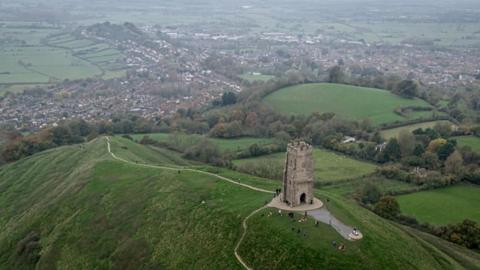 An aerial image shows Glastonbury Tor with the town in the background, on a cloudy summer's day. Around a dozen people can be seen on the hill near the Tor.