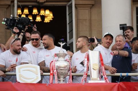 Hull KR players, wearing white shirts are standing on the balcony of Hull City Hall with three trophies in front of them