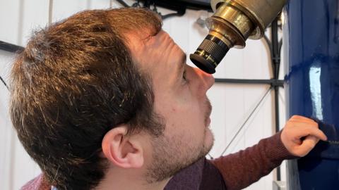 Andrew Marshall-Lee has short brown hair. He is looking up into a golden telescope. He is wearing a maroon sweater and has his left hand on the blue base of the telescope. 