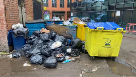Fly-tipped waste in Coventry city centre. A pile of black bin bags are piled on top of each other with large waste bins overflowing.