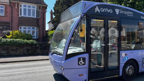A light purple bus, with 'arriva' and 'Shropshire Council' written on it in white writing. It's driving along a road with a red brick house visible on the other side of the street. The sky is bright blue and there's strong sunshine. 