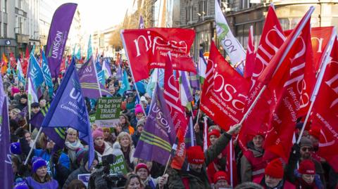 Hundreds of people from different trade unions stand in a Belfast street waving red, blue, purple and green flags