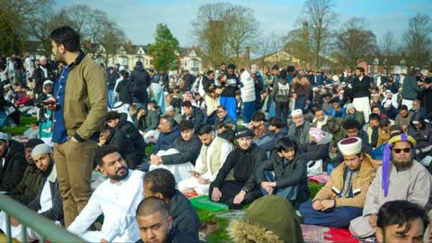 A crowd of men standing or sitting in a park, with trees and houses in the background. Many of them are wearing religious clothing.