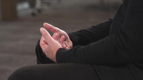 The hands of a man sitting on on a chair, wearing a black jumper and trousers