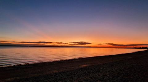 A beautiful sunset over a beach. The sky is an orange glow in the distance with the sky turning more blue and dark above. The sea can be seen lit up by the sunset while the beach os covered in darkness.