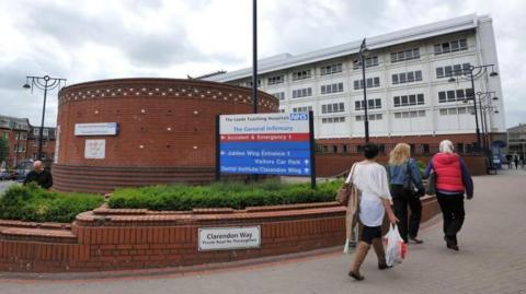 Three women walk past the sign for a Leeds General Infirmary. It is a red brick and white building