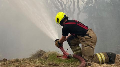 A firefighter wearing a fluorescent yellow helmet and uniform directs a jet of water from a red hose. He kneels on a grassy mound with trees in the background and is surrounded by hazy smoke.