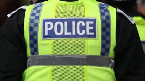 A stock image of the back of a police officer wearing a yellow hi-vis vest with blue and grey checks running down the sides. Police is written in the middle of the vest in white letters in a blue box. The officer is also wearing a black long-sleeved top underneath.