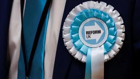 Close-up photo of a blue and white Reform UK rosette worn by someone wearing a dark suit. 