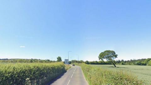 Google Maps image of the Saltburn Lane looking towards the A174 roundabout. The road is single-carriageway in each direction in between fields. A hedge grows very close to the road's edge on both sides.