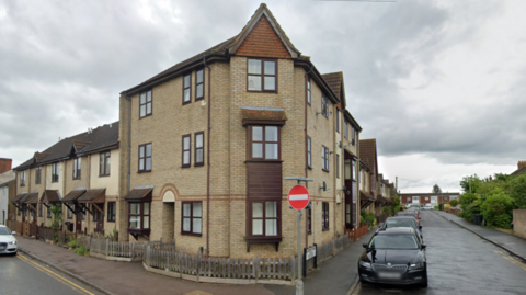 A block of brown-brick flats on a corner where two streets meet. Rows of houses lead to the block. Cars are parked in each street.