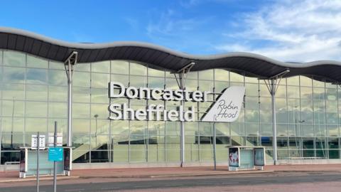 Against a backdrop of a blue sky, a wide glass fronted building with a wavy roof is at the side of a road. There are two bus stops spaced out in front of the building. Large white letters on the front of the building read Doncaster Sheffield. A white sign to the right of them reads Robin Hood.