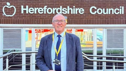 A man wearing a grey suit and a yellow lanyard is standing outside a building which has white lettering indicating it is a Herefordshire Council office.