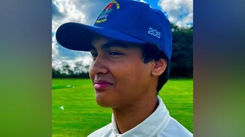 Absi Ahmed smiling on a cricket pitch looking at the camera side on. He is wearing a white cricket top and a blue cricket cap. There are clouds in the otherwise blue sky. 
