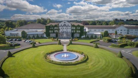 An aerial shot of a three-storey white building with ivy over the front of the building. On either side is a car park, and the building sits in front of a circular green patch of grass with a fountain in the middle.