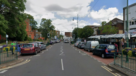 Cars are parked on either side of the high street with people on both sides of the road. There are more shops on the right hand side of the image.