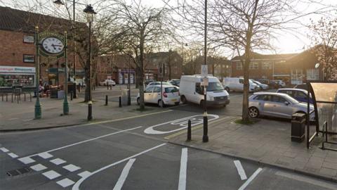 Google street view of Cottingham's Market Place car park with a large green clock with the name Cottingham on it and a number of vehicles parked in bays in a smart-looking square.