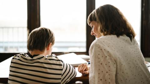 A boy and a woman are sat at a table with their backs to us. The boy is wearing a black and white striped top and looking at a notebook. The woman is wearing a white top and looking down at the boy's notebook.