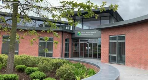 Front of Acute Mental Health Centre. There is a flower bed with shrubs and trees. The building has brown bricks and glass doors.