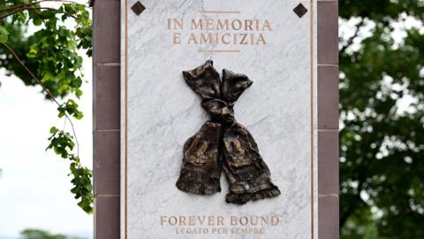A memorial of two scarves knotted together on a marble background. Gold text reads: In memoria e amicizia. Forever Bound. Legato Per Sempre