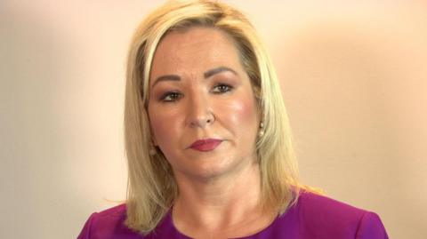 Michelle O'Neill has blond hair down to her shoulders, brown eyes and is wearing a purple top 