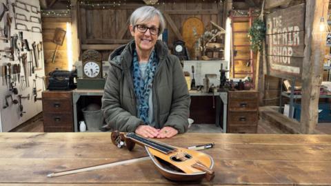 A woman in a jacket and scarf sat behind a wooden table. A yellow lyra and its bow are resting on the table. She is sat on the set of The Repair Shop, with several antiques and tools visible in the background.
