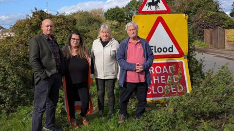 Four people stood on the side of the road. They are looking into the camera with a large bush behind them. To one side is a large "flood" sign that also says "road ahead closed".