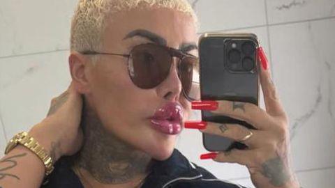 Jordan Parke seen with closely cropped bleached blonde hair takes a mirror selfie while holding a smartphone. They are wearing large dark sunglasses, a black zip-up top and a gold watch. Long, bright red manicured nails are visible as they hold the phone, and tattoos can be seen on their neck and hand. The background appears to be a tiled bathroom wall.