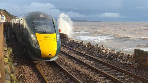 A GWR train passes a section of crumbled wall. It is next to a churned up sea.