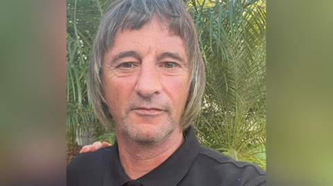 Ian Woolfenden wears a black polo shirt. He has short grey stubble and chin-length grey/brown hair with a short fringe. He is standing outside.