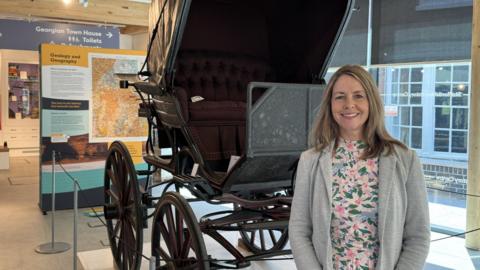 Hayley Coles, a woman wearing a floral top and a grey jacket, is standing next to a black carriage. In the background is a museum information board and a barrier to protect the carriage.