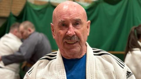 Harry McGuigan is an older man with a greying moustache. He is bald and is wearing a white Judo jacket with black stripes on the shoulders. He's standing in a leisure centre hall and behind him are other people taking part in Judo grappling.  