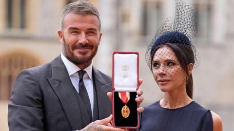 Sir David and Lady Beckham pose for the cameras outside Windsor Castle with Sir David's honour. He is wearing a grey suit, white shirt and grey tie and she is in a navy sleeveless dress and pillarbox hat with netting.