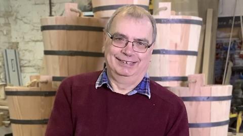 Allan Paulus wearing a red jumper and glasses. He's holding a bucket and is stood in front of a pile of buckets in his workshop