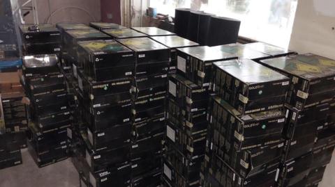 Several piles of black boxes stacked up in a room. The boxes have green and yellow details on one side as well as white writing.