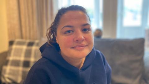 Vicky Prussia, wearing a blue hooded top, looking at the camera while sitting in a living room