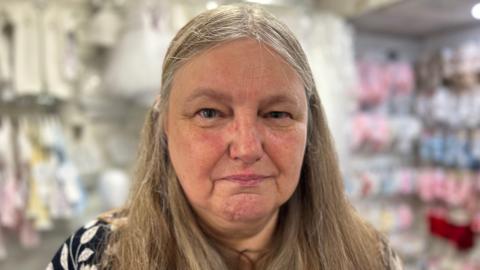 A lady with grey hair and a black and white top looking at the camera with a glum face. There are rows of baby clothes in the background.