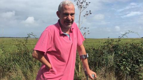 Steven Lacey standing against a countryside backdrop. Steven is standing with a crutch
