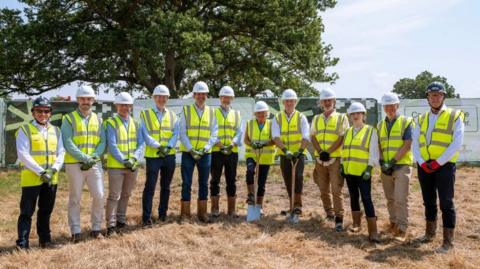 Twelve people, all in high vis jackets and hard hats, pose for a photo in a line. Two hold spades, while they all stand on brown grass, with a big oak tree in the background.