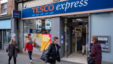 Out the front of a Tesco express store, with several shoppers strolling passed.