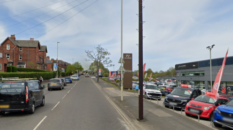 Cars drive away from the camera along a main road. On the right is a car sales forecourt. On the left are some large redbrick houses in the distance.