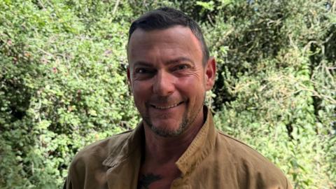 James Ackroyd-Cooper is smiling at the camera in a photo taken on a sunny day in front of some trees. He has short, brown hair and facial hair. He is wearing a tan-coloured jacket.