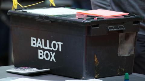 A black ballot box at a vote count. The words ballot box are written in white block capitals on the side of the box. A large calculator is next to the box, which is on a grey table. Pieces of orange paper are on top of the box.
