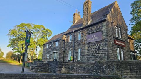 A pub building, built from heritage stones. It has maroon signs with gold writing identifying it as 'The Dickie Bird'. It seems to be in a semi rural setting and is surrounded by trees. 
