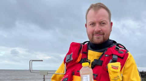 An environment agency water tester stands on Sheringham beach showing a sample of water in a bottle to the camera. He is wearing a yellow waterproof suit and a red gillet. 